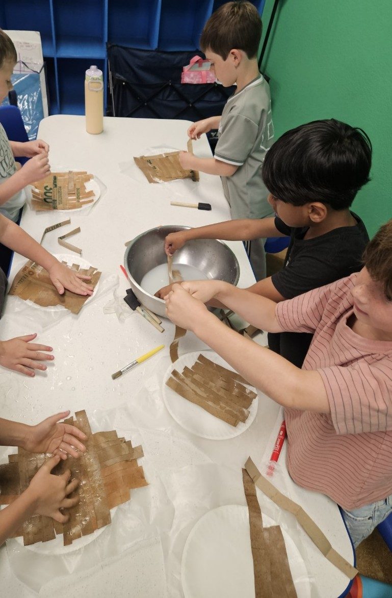 Children weaving strips of reed into papyrus sheets around a table